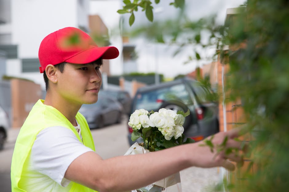 A delivery person wearing a red cap and a neon yellow vest is placing a floral gift consisting of white roses and green foliage into a flower box on a brick wall outdoors. The bouquet is wrapped in wh