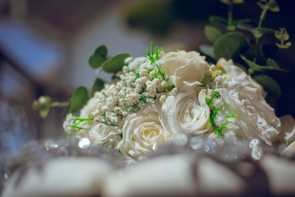 A close-up of a wedding floral arrangement featuring white roses with soft, layered petals, complemented by small white baby's breath flowers and sprigs of green eucalyptus leaves. The bouquet is rest