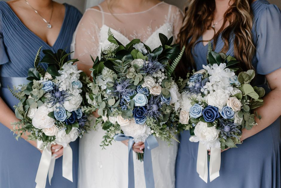 A close-up image featuring three bridal bouquets, each composed of white and light blue roses, white hydrangeas, and sprigs of blue thistles, accented with various shades of green foliage including eu