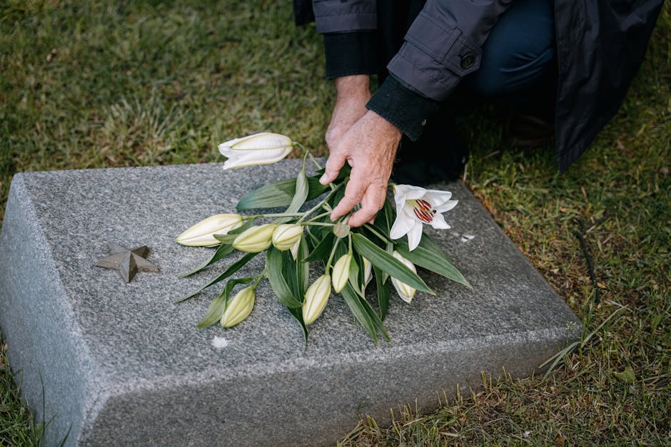 A person placing a bouquet of white lilies and lilies of the valley with green leaves onto a polished grey granite memorial stone, which has a small metal star embedded on its surface, during a floral
