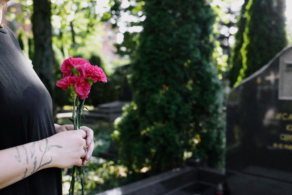 A person holding a small bouquet of bright pink carnations with ruffled petals and green stems, wrapped with a clear plastic cover. The individual, wearing a black top, has a visible tattoo on their w