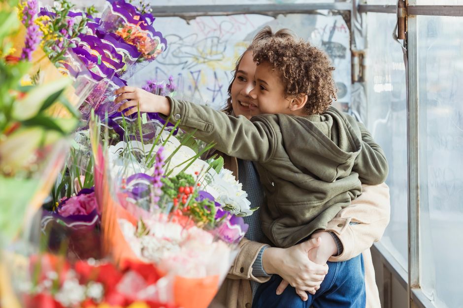 A young child with curly hair and a grey hoodie is being held by an adult, possibly a parent, inside a flower shop. The child is reaching out happily towards a vibrant display of fresh floral bouquets