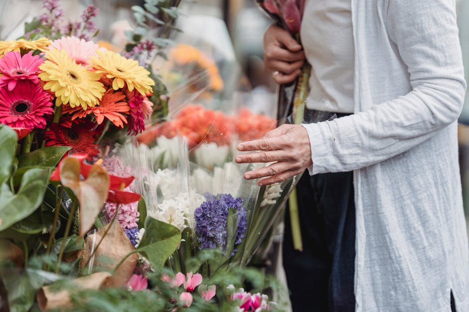 A person wearing a white long-sleeve shirt and dark trousers is selecting a bouquet of fresh flowers at a flower market. The arrangement features vibrant, multi-colored blooms including bright pink ge